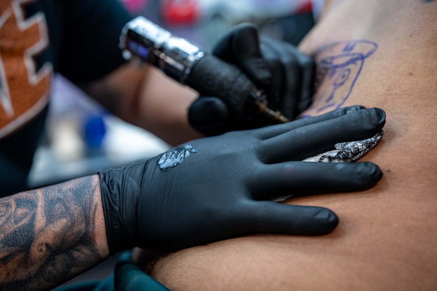 Ein Master in schwarzen Handschuhen arbeitet mit der Tätowiermaschine auf Haut mit blauem Stencil, Sekundärgel auf geöffneter Haut während der Arbeit